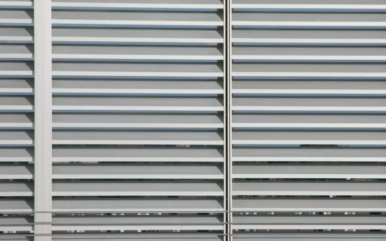 Zdjęcie przedstawia ogrodzenie żaluzjowe wykonane z metalowych, poziomych listew. Listwy te są ustawione pod kątem, co przypomina żaluzje okienne. Konstrukcja składa się z dwóch głównych pionowych słupków, do których przymocowane są listwy. Ogrodzenie to zapewnia zarówno prywatność, jak i przepuszczalność powietrza, dzięki szczelinom między listwami. Całość ma nowoczesny wygląd i jest utrzymana w jasnej, metalicznej kolorystyce. W tle można dostrzec zarys innych budynków i elementów krajobrazu, które są widoczne przez szczeliny ogrodzenia.