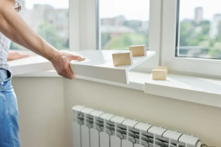 Obraz przedstawia osobę, prawdopodobnie mężczyznę, który montuje parapet wewnętrzny przy oknie. Mężczyzna ubrany jest w niebieskie dżinsy i koszulę w kratę, a jego dłoń trzyma białą płytę parapetu. W tle widoczne jest duże okno, przez które widać rozmyty krajobraz miasta z budynkami oraz zielenią. Na grzejniku pod oknem znajdują się drewniane klocki, które mogą być używane do podtrzymania lub poziomowania parapetu podczas montażu. Całość tworzy wrażenie pracy remontowej lub wykończeniowej wewnątrz mieszkania, co może wywoływać uczucie porządku, nowości i dbałości o detale w domowym otoczeniu.