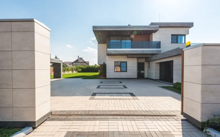 Obrazek przedstawia nowoczesny dom z zadaszeniem nad wejściem. Budynek jest dwupiętrowy, wykonany z jasnych materiałów, prawdopodobnie betonu lub kamienia. Zadaszenie nad wejściem jest prostokątne, wspierane na bocznych ścianach, co tworzy minimalistyczny i elegancki wygląd. ### Elementy widoczne na obrazie: 1. **Budynek**: - Dwupiętrowy, nowoczesny dom z płaskim dachem. - Ściany są pokryte jasnym materiałem, prawdopodobnie kamieniem lub betonem. - Duże okna, które wpuszczają dużo naturalnego światła. - Balkon na pierwszym piętrze z przezroczystą balustradą. 2. **Podjazd**: - Szeroki, wykonany z kostki brukowej o jasnym kolorze, z ciemniejszymi wzorami. - Podjazd jest czysty i dobrze utrzymany, prowadzi bezpośrednio do wejścia do domu. 3. **Otoczenie**: - Z lewej strony widoczny jest fragment zielonego trawnika. - W tle, za domem, widać inne budynki i drzewa, co sugeruje, że dom znajduje się w spokojnej, prawdopodobnie podmiejskiej okolicy. Ogólnie, obrazek przedstawia elegancki, nowoczesny dom z przestronnym podjazdem, otoczony zielenią, co tworzy przyjemne i estetyczne otoczenie.