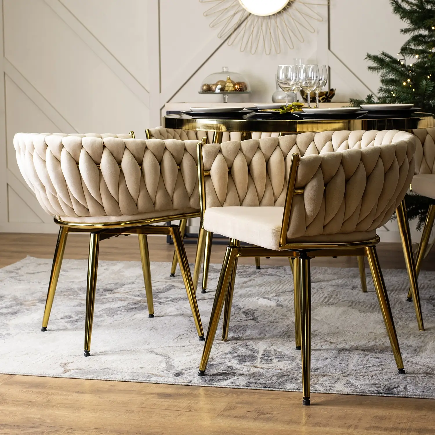 Zdjęcie przedstawia elegancką aranżację wnętrza z nowoczesnymi, luksusowymi krzesłami o złotych nogach. Krzesła mają zaokrąglone oparcia i są obite beżowym, pikowanym materiałem, który wygląda na miękki i wygodny. Tkanina ma charakterystyczny, elegancki wzór przypominający plecionkę. Krzesła stoją na podłodze z drewnianych paneli, na której znajduje się jasny dywan z subtelnym wzorem. W tle widać część stołu z eleganckimi naczyniami, kieliszkami oraz dekoracją świąteczną, co sugeruje, że jest to jadalnia przygotowana na specjalną okazję. Na ścianie wisi okrągłe lustro z dekoracyjną ramą, a w rogu pomieszczenia znajduje się udekorowana choinka, co dodaje atmosfery świątecznej.