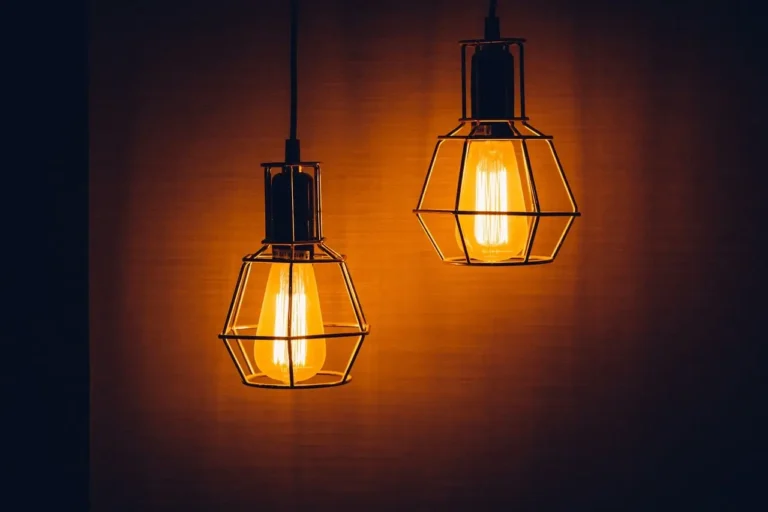 Na zdjęciu widzimy dwie wiszące lampy w stylu industrialnym, które emitują ciepłe, złociste światło. Lampy mają metalowe klosze w kształcie klatek, co nadaje im charakter vintage lub loftowy. Żarówki w stylu retro (typu Edison) podkreślają surowy, ale przytulny charakter oświetlenia. Tło jest jednolite, w ciemnym, ciepłym odcieniu, co dodatkowo wzmacnia atmosferę przytulności i ciepła.