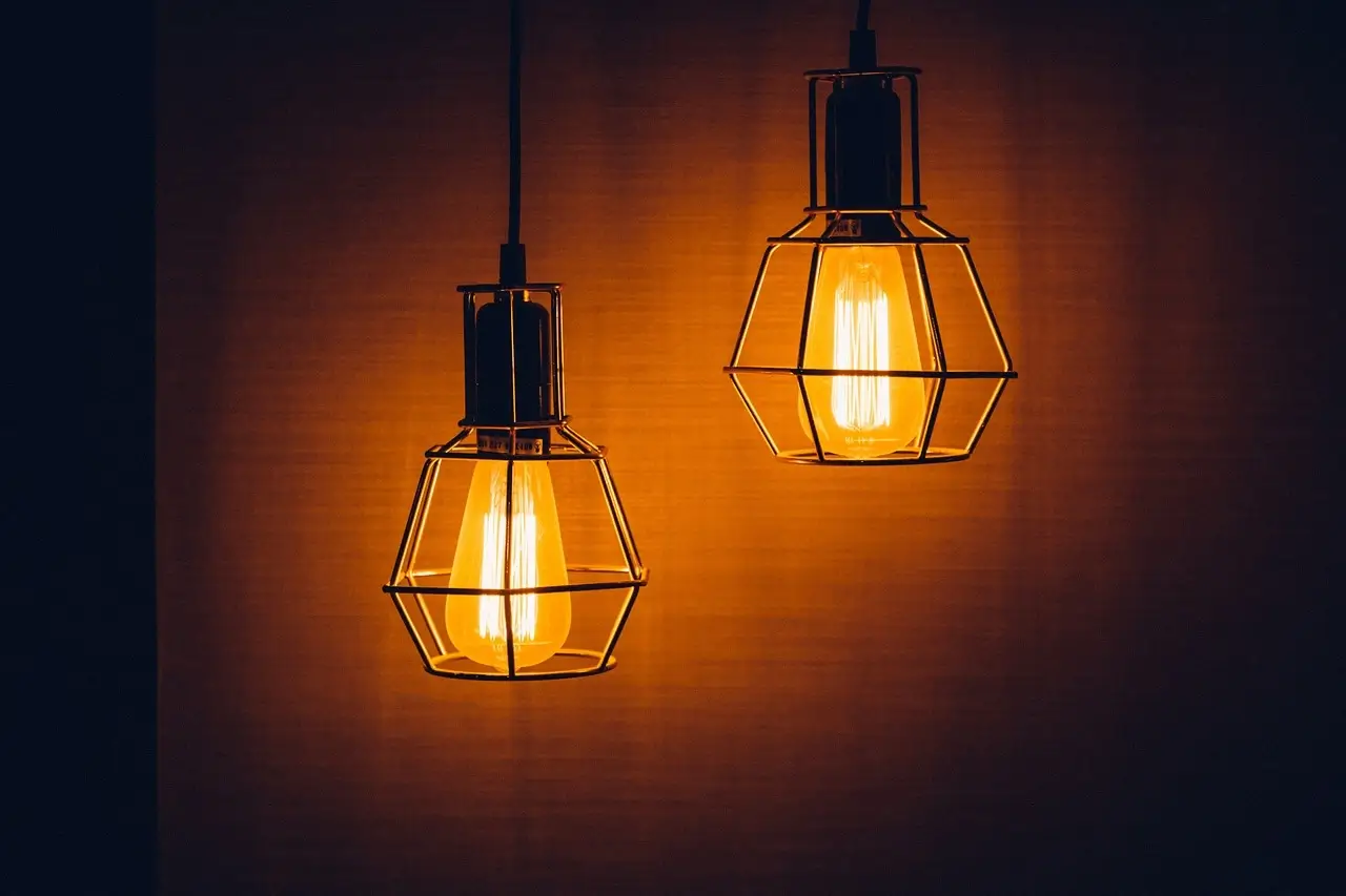 Na zdjęciu widzimy dwie wiszące lampy w stylu industrialnym, które emitują ciepłe, złociste światło. Lampy mają metalowe klosze w kształcie klatek, co nadaje im charakter vintage lub loftowy. Żarówki w stylu retro (typu Edison) podkreślają surowy, ale przytulny charakter oświetlenia. Tło jest jednolite, w ciemnym, ciepłym odcieniu, co dodatkowo wzmacnia atmosferę przytulności i ciepła.