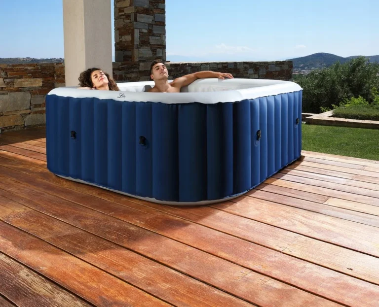 Na zdjęciu widać dwoje ludzi relaksujących się w nadmuchiwanym jacuzzi umieszczonym na drewnianym tarasie na zewnątrz. Jacuzzi ma prostokątny kształt z zaokrąglonymi rogami, ciemnoniebieskie ściany zewnętrzne oraz biały środek. Niebieskie ściany mają pionowe prążkowania dla stabilności i wyposażone są w czarne zawory, prawdopodobnie do pompowania lub spuszczania wody. W środku siedzą mężczyzna i kobieta. Kobieta po lewej stronie ma kręcone brązowe włosy i patrzy w górę z wyrazem spokoju na twarzy. Mężczyzna po prawej stronie ma krótkie brązowe włosy, leży wygodnie z jedną ręką opartą na krawędzi jacuzzi, również wyglądając na zrelaksowanego. Otoczenie to malowniczy taras na świeżym powietrzu. Pod jacuzzi znajduje się drewniana podłoga z desek o naturalnym wykończeniu, lekko postarzałych od słońca. Za jacuzzi widać niski mur z dekoracyjnych cegieł, a w tle rozciąga się widok na trawnik, krzewy i wzgórza. Niebo jest jasnoniebieskie, co sugeruje słoneczny, pogodny dzień.