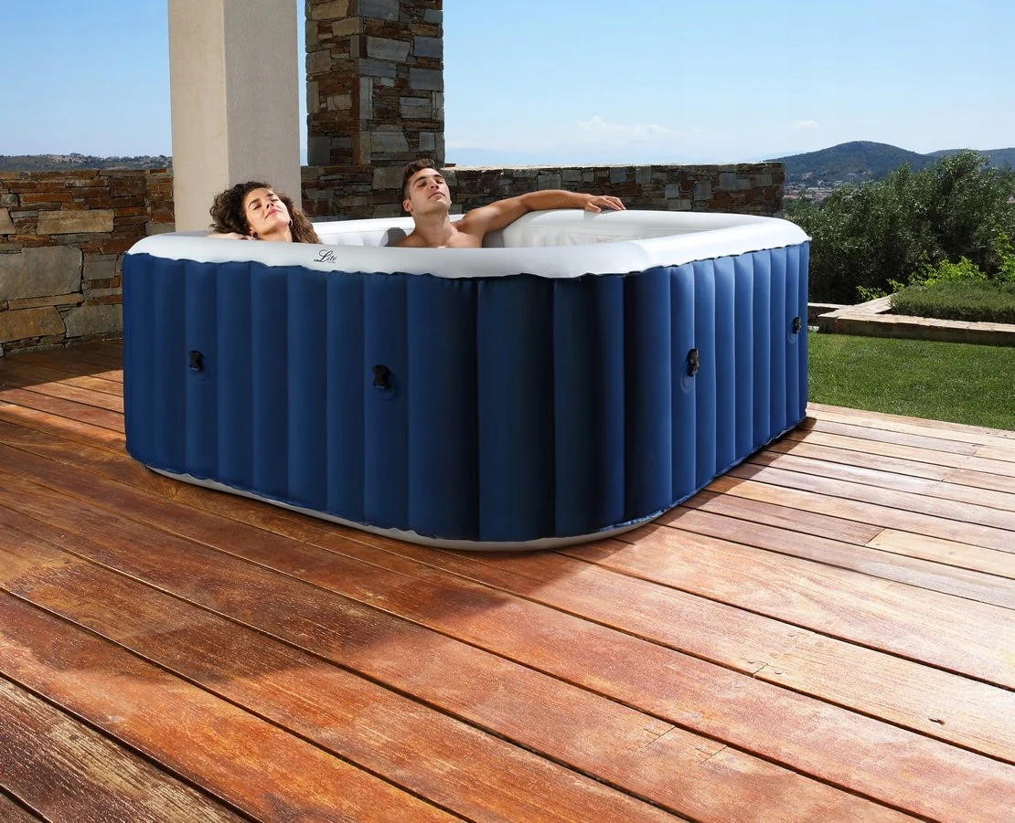 Na zdjęciu widać dwoje ludzi relaksujących się w nadmuchiwanym jacuzzi umieszczonym na drewnianym tarasie na zewnątrz. Jacuzzi ma prostokątny kształt z zaokrąglonymi rogami, ciemnoniebieskie ściany zewnętrzne oraz biały środek. Niebieskie ściany mają pionowe prążkowania dla stabilności i wyposażone są w czarne zawory, prawdopodobnie do pompowania lub spuszczania wody. W środku siedzą mężczyzna i kobieta. Kobieta po lewej stronie ma kręcone brązowe włosy i patrzy w górę z wyrazem spokoju na twarzy. Mężczyzna po prawej stronie ma krótkie brązowe włosy, leży wygodnie z jedną ręką opartą na krawędzi jacuzzi, również wyglądając na zrelaksowanego. Otoczenie to malowniczy taras na świeżym powietrzu. Pod jacuzzi znajduje się drewniana podłoga z desek o naturalnym wykończeniu, lekko postarzałych od słońca. Za jacuzzi widać niski mur z dekoracyjnych cegieł, a w tle rozciąga się widok na trawnik, krzewy i wzgórza. Niebo jest jasnoniebieskie, co sugeruje słoneczny, pogodny dzień.