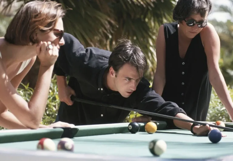Zdjęcie przedstawia troje młodych dorosłych grających w bilard na świeżym powietrzu. Szczegóły obrazu: • Mężczyzna w czarnej koszuli jest w centrum uwagi – pochyla się nad stołem bilardowym, koncentrując się na uderzeniu bilą kijem bilardowym. Jego wyraz twarzy zdradza skupienie i determinację. • Dwie kobiety obserwują grę. • Kobieta po lewej stronie, ubrana na biało, opiera się o stół, trzymając dłoń pod brodą, co sugeruje, że jest zainteresowana i może analizować ruch gracza. • Kobieta po prawej, ubrana na czarno i nosząca ciemne okulary, również przygląda się grze, stojąc nieco dalej. • Stół bilardowy jest pokryty zielonym suknem, a kilka bil jest rozsianych po jego powierzchni, w tym kula 8 oraz żółta i czarna bila. • Tło jest rozmyte, ale widać palmy i jasne światło słoneczne, co sugeruje, że scena dzieje się w ciepłym klimacie lub letnim okresie. Atmosfera i przekaz: Zdjęcie oddaje relaksującą i towarzyską atmosferę. Trójka znajomych spędza czas na świeżym powietrzu, grając w bilard, co sugeruje beztroską zabawę i rywalizację w przyjaznym gronie. Naturalne światło i otoczenie palmowe dodają wrażenia wakacyjnej swobody.