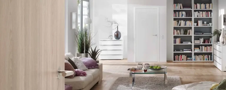 Zdjęcie przedstawia elegancki, nowoczesny salon widziany przez uchylone, jasnobrązowe drewniane drzwi z metalową klamką. Wnętrze jest przestronne, jasne i urządzone w minimalistycznym stylu, z dominacją bieli, beżu i naturalnych elementów. Po lewej stronie znajduje się jasna, kremowa sofa ozdobiona poduszkami w różnych odcieniach fioletu i bieli. Obok sofy stoi wysoka roślina doniczkowa oraz biała lampa podłogowa. Na środku pokoju umieszczono szklany stolik kawowy na metalowych nogach, na którym leży kilka dekoracyjnych przedmiotów: szklany dzbanek, filiżanka herbaty oraz misa z zielonymi jabłkami. Stolik stoi na miękkim, jasnym dywanie, który kontrastuje z drewnianą podłogą. Po prawej stronie znajduje się duża, biała biblioteczka wypełniona książkami ułożonymi zarówno pionowo, jak i poziomo, a także ozdobnymi wazonami i figurkami. Obok biblioteczki stoi biała komoda z dekoracyjnymi wazonami i rośliną. W głębi pokoju widać białe drzwi oraz niewielką, białą komodę z ozdobnymi wazonami i suchą gałązką. Na ścianie wiszą proste, minimalistyczne obrazy. Całe wnętrze jest dobrze doświetlone naturalnym światłem wpadającym przez duże okno, co podkreśla jego przytulny, harmonijny i elegancki charakter.